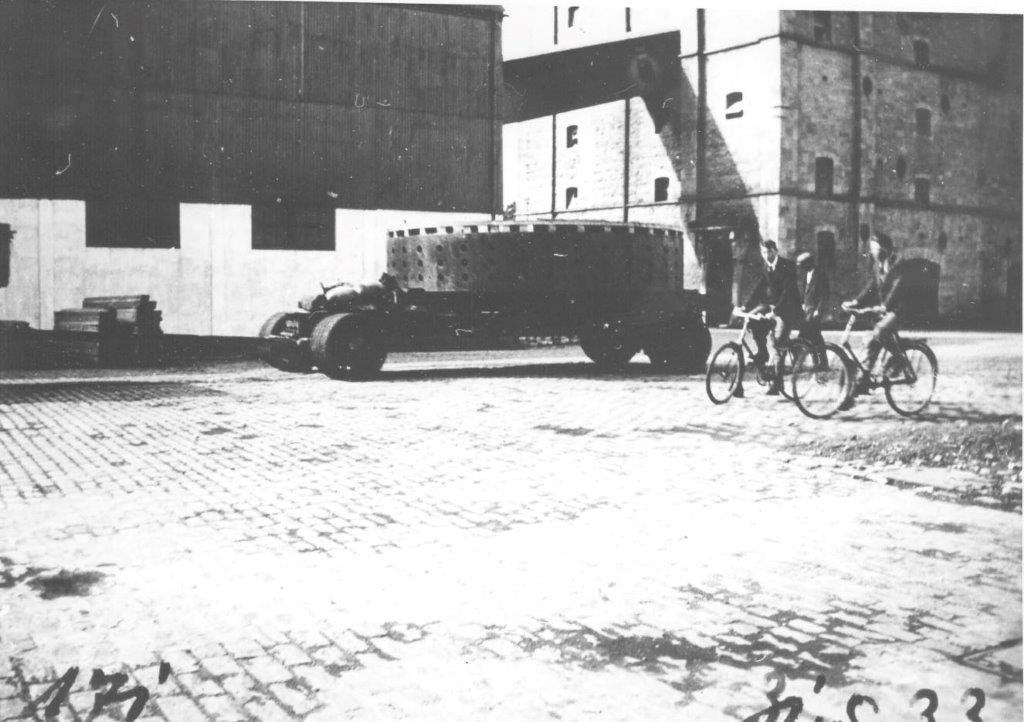 Black and white photograph of a large cylindrical generator component mounted on a transport vehicle crossing a cobbled city street, with two cyclists riding nearby and industrial buildings in the background.