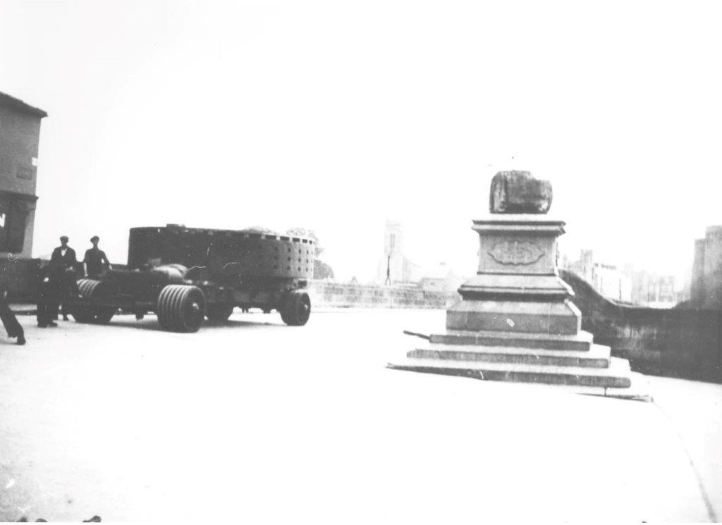 Black-and-white photograph showing a generator component being transported through Limerick city on a multi-wheeled carriage, passing the Treaty Stone on its stone plinth beside the river.