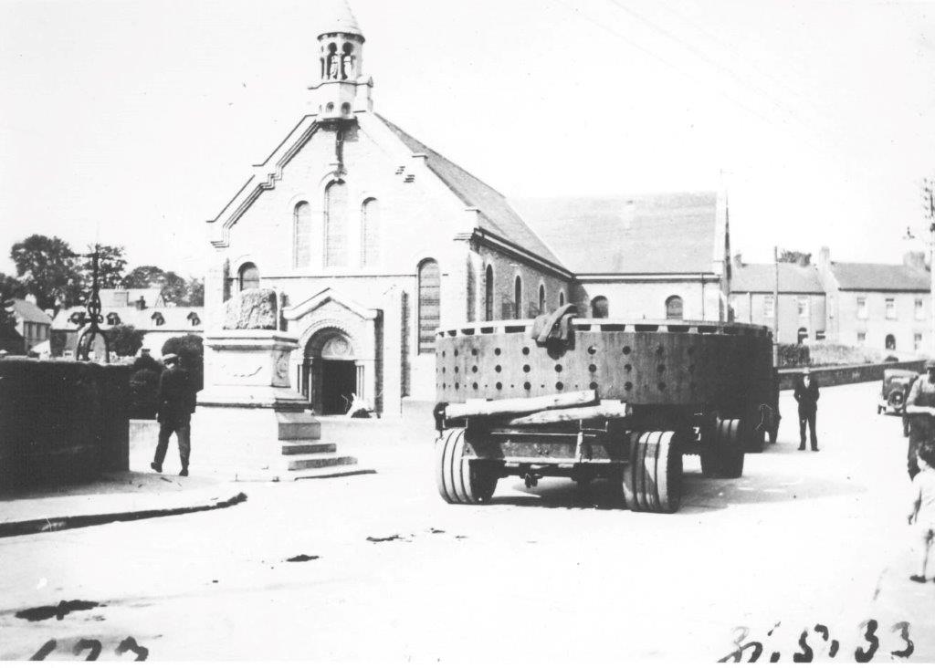 Black and white photograph showing a large cylindrical generator component on a multi-wheeled transport carriage passing the Treaty Stone and a church building, with several people standing nearby on a town street.