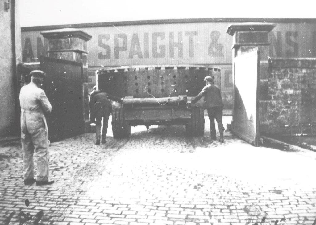 Black-and-white photograph showing a generator component being carefully manoeuvred through a gated entrance between industrial buildings, with workers guiding the transport vehicle and an onlooker standing nearby.