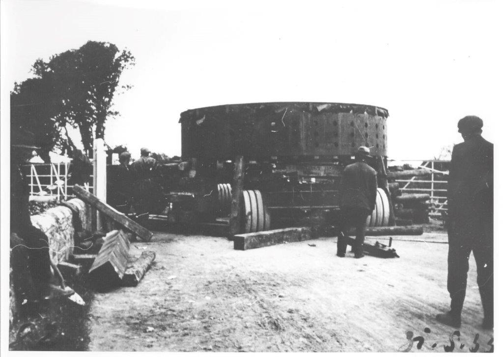 Black-and-white photograph showing a large generator component resting on a multi-wheeled transport carriage, with onlookers and workers positioned around it beside a country road and gate.