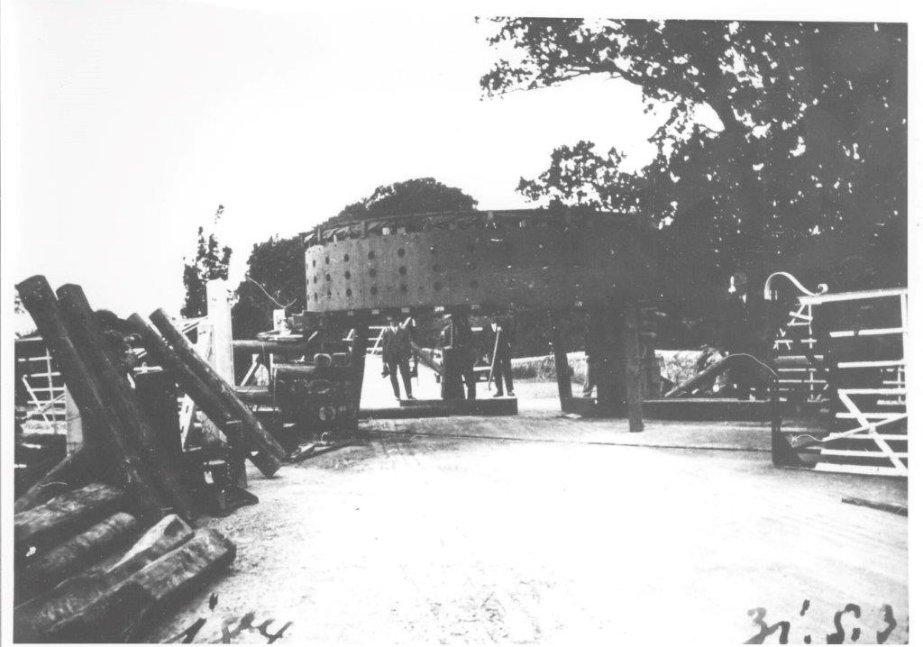 Black-and-white photograph showing a large generator component on a transport carriage passing between gates on a rural roadway, with workers nearby and stacked timber in the foreground.