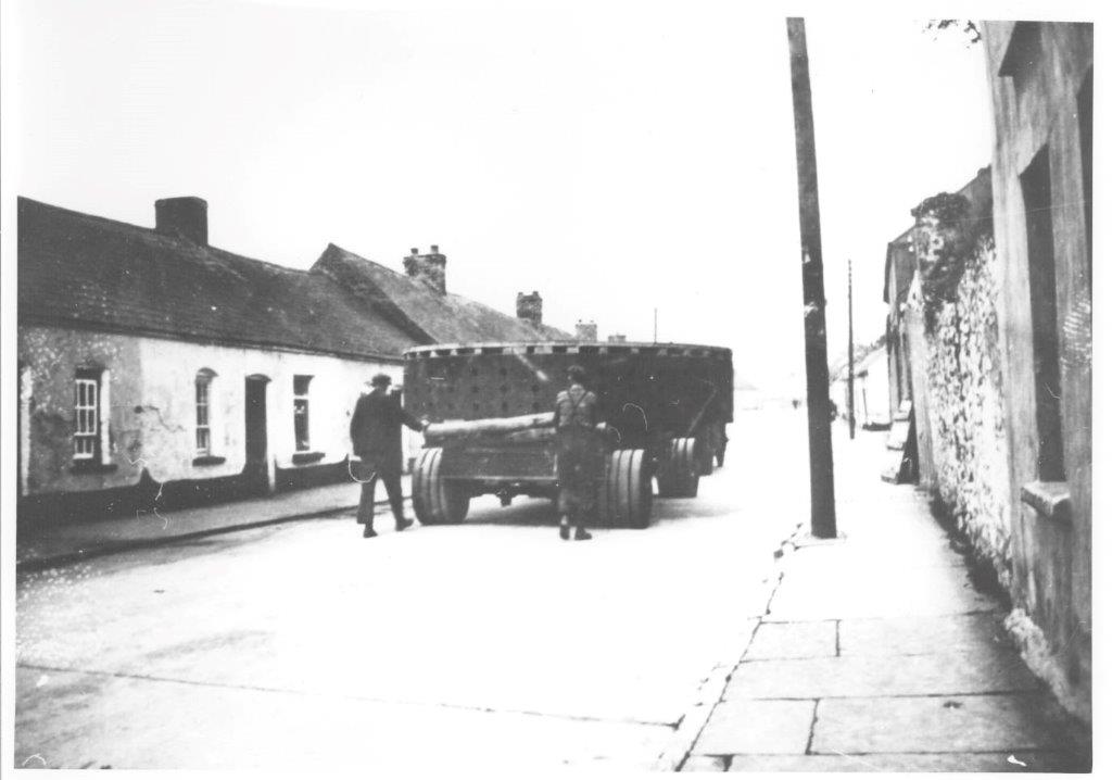 Black-and-white photograph showing a large circular generator component being transported through a village street on a heavy wheeled platform, with people guiding the load between closely spaced buildings.