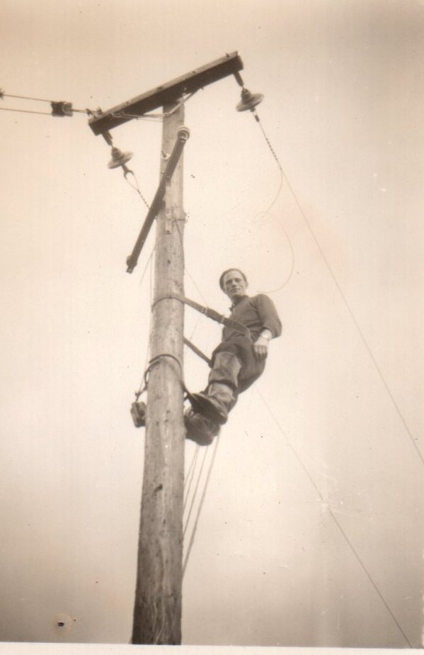 Tim Slevin climbing a wooden electricity pole using pole-climbing equipment, with overhead power lines and insulators visible at the top of the pole.