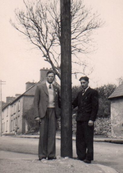 Tim Slevin standing beside an electricity pole on a village street with another man, with houses and a tree visible in the background.