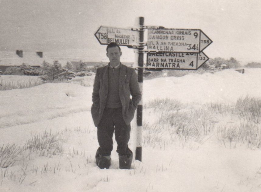 Tim Slevin standing in deep snow beside a roadside signpost showing directions and distances to nearby locations.