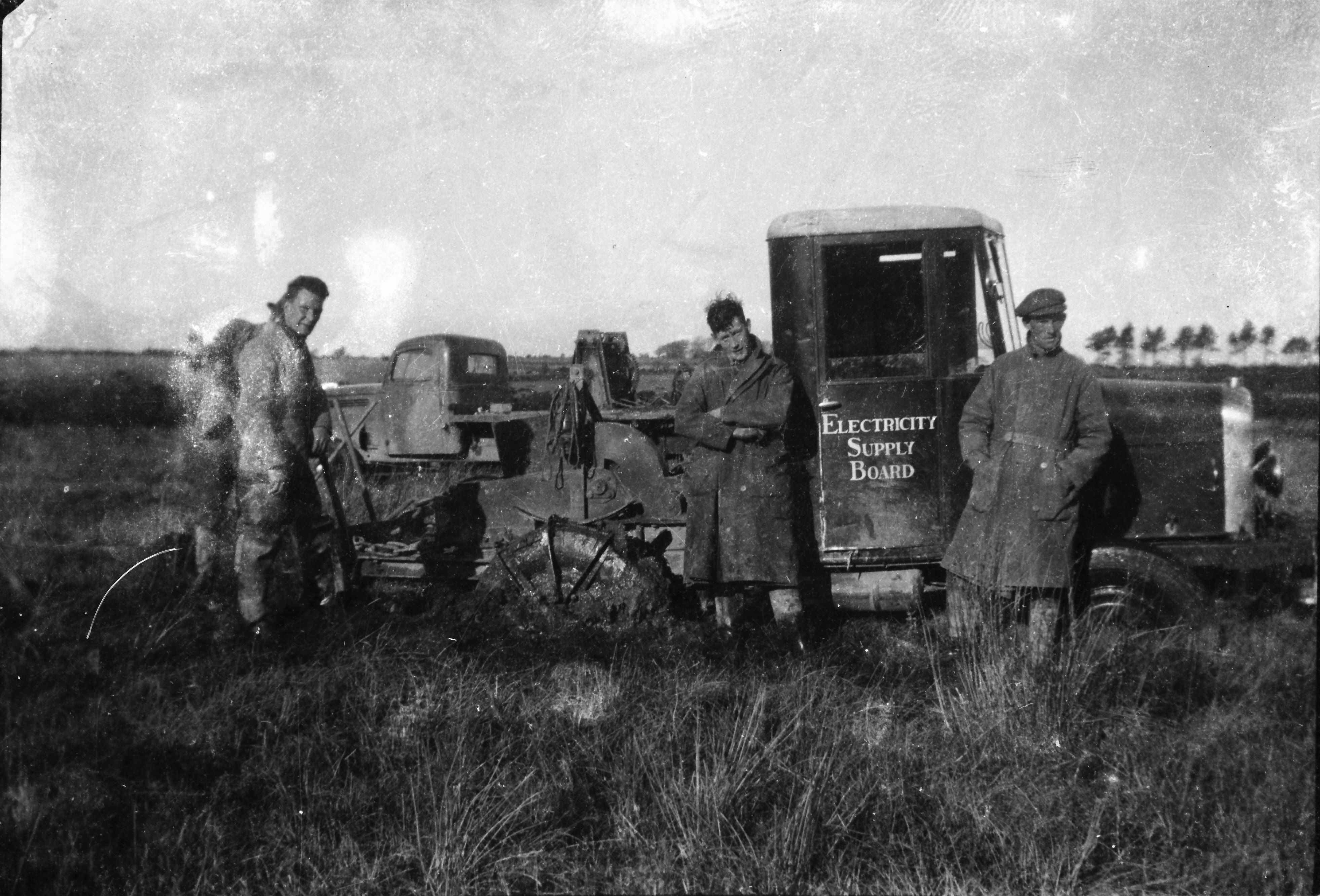 Three ESB workers standing beside a line construction vehicle fitted with excavation or pole-setting equipment in a grassy field.