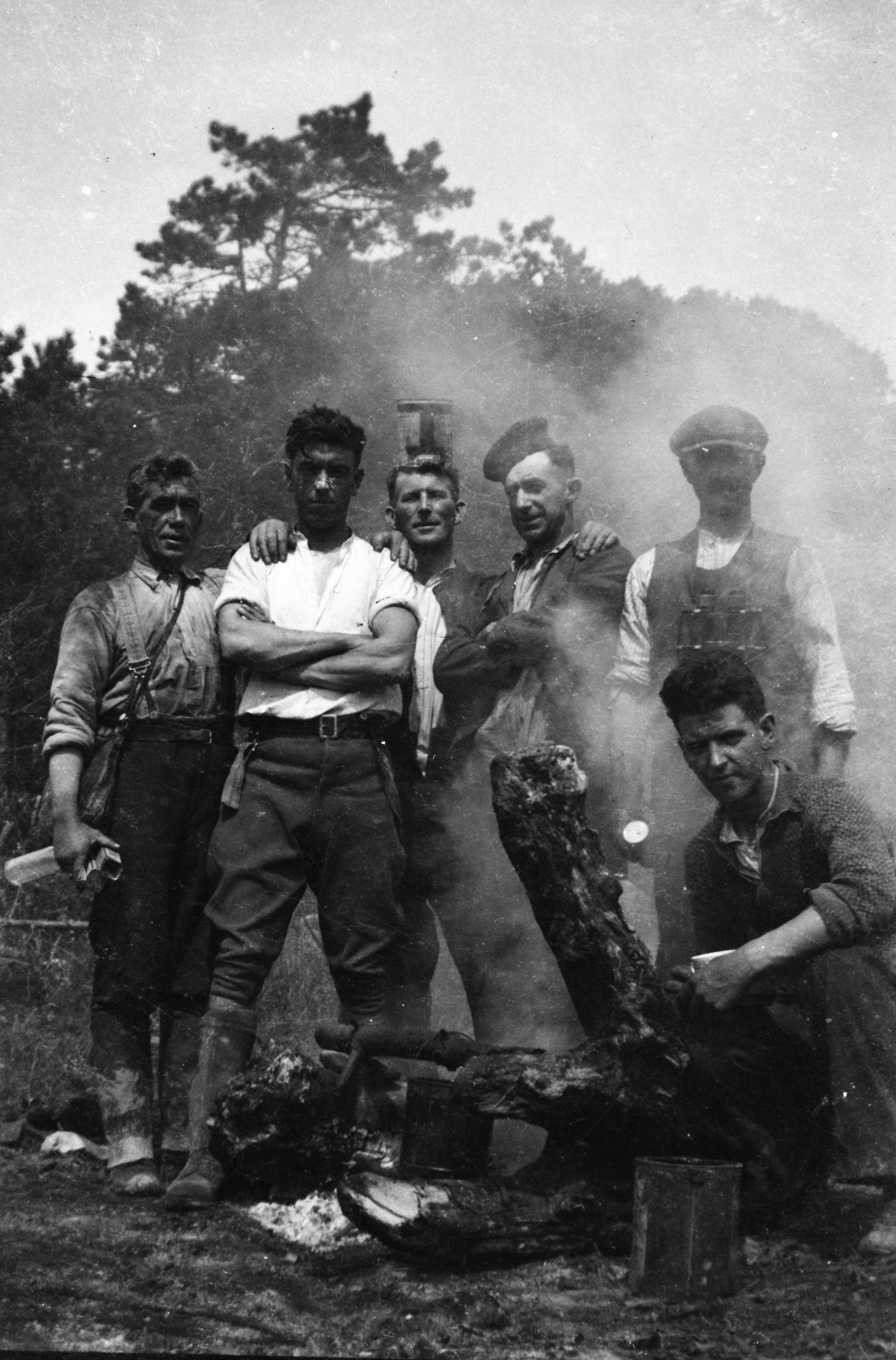 Black-and-white photograph of Jerry Corbett and five of his ESB colleagues on the rural electrification scheme standing and crouching around a campfire at an outdoor work site, with cooking equipment visible in the foreground.