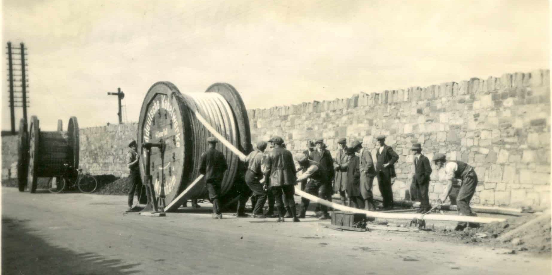 Black-and-white photograph showing workers laying electricity distribution cables along a roadside. A large cable drum is being unwound while a group of workers guide the cable beside a stone wall.