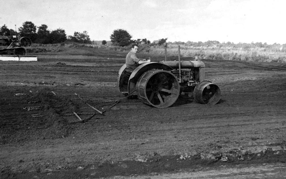 Tractor operating on a prepared bog during peat harvesting, with additional machinery visible in the background.