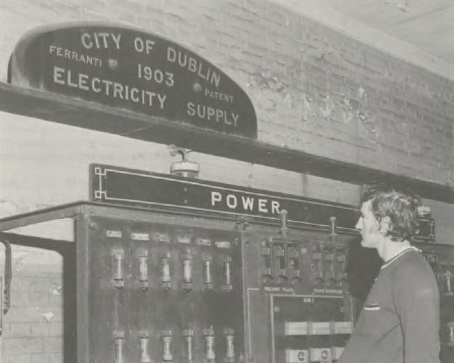 Person inspecting an early electricity control panel beneath a sign reading “City of Dublin Electricity Supply”.