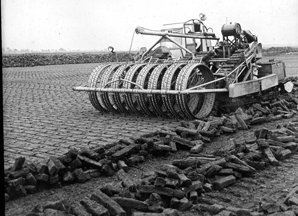 Mechanical peat-harvesting machine operating on a bog, with rows of cut peat in the foreground and patterned peat fields in the background.