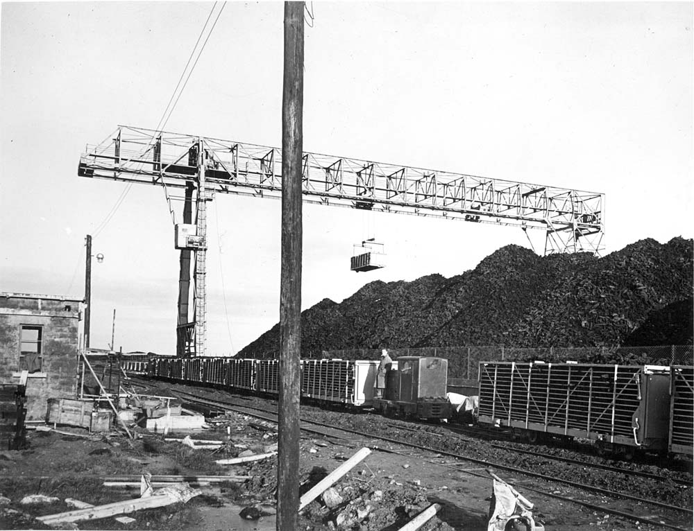 Overhead gantry crane operating above railway wagons at a peat storage yard, with large peat stockpiles in the background.