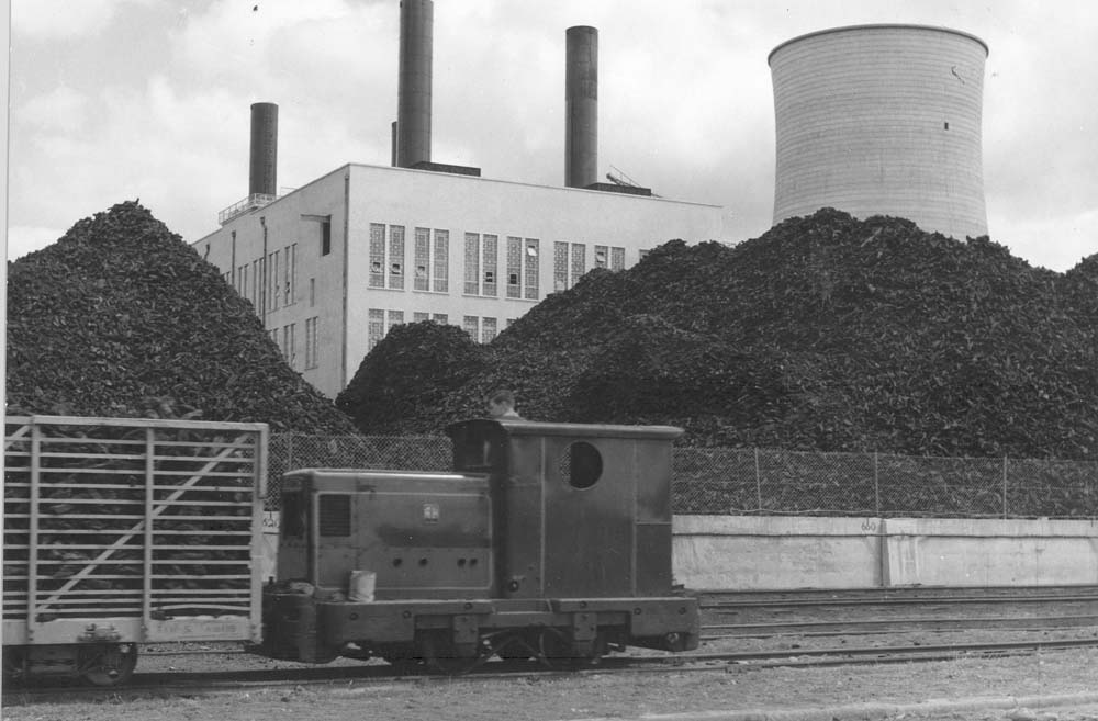 Small locomotive and rail wagon on industrial tracks in front of large peat stockpiles and a power station.