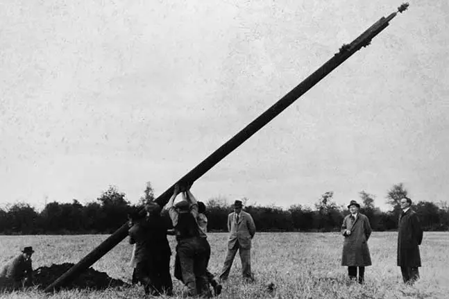 Photograph of an electricity pole being installed in a rural Irish community during the Rural Electrification Scheme. There are three men in suits watching the pol being erected.