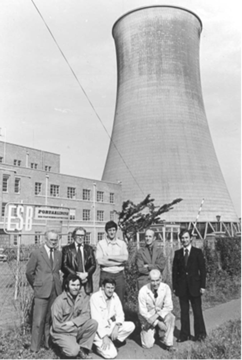 A group of 8 men in front of Portarlington power station chimney with an ESB logo behind them.