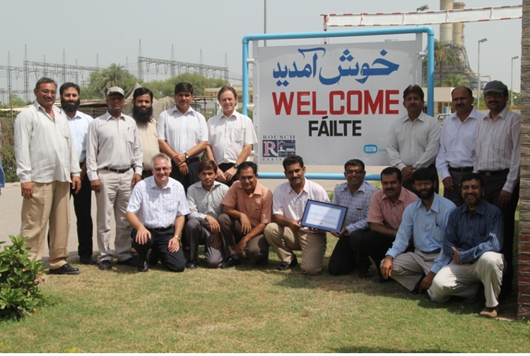 Group photograph taken outdoors beside a sign displaying “WELCOME” and “FÁILTE”, with industrial infrastructure visible in the background.