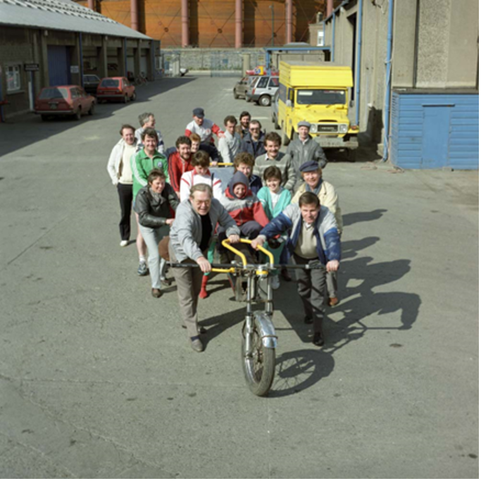 Group of people pushing and riding a large multi-person bicycle through an industrial yard, with parked vehicles and warehouse buildings in the background.