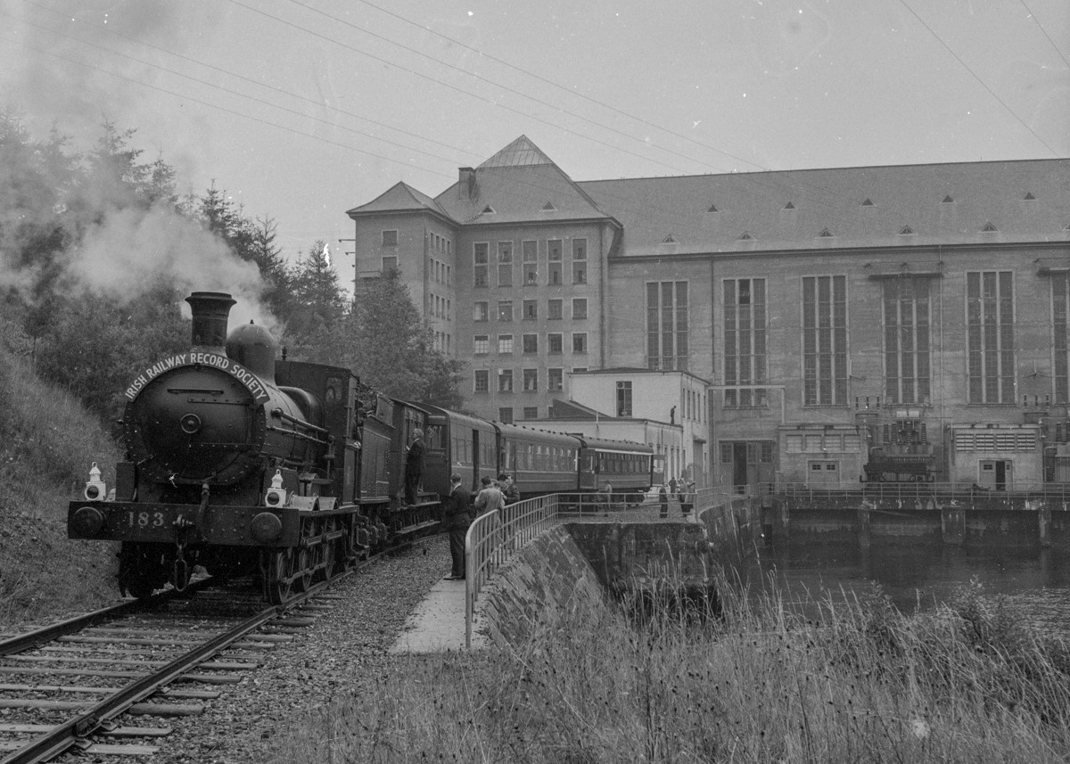 Black-and-white photograph showing a steam excursion train stopped beside the Ardnacrusha hydroelectric power station, with passenger carriages and visitors visible alongside the track.