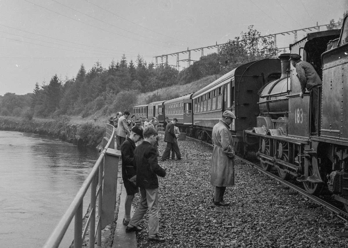 Black-and-white photograph showing a steam train carrying visitors along the railway beside a waterway near Ardnacrusha power station.