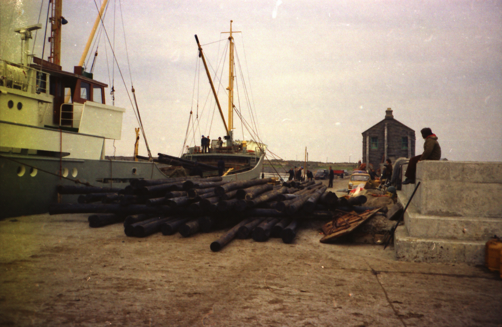 Colour photograph of wooden electricity poles being unloaded from a ship at a harbour, with onlookers watching in the background.