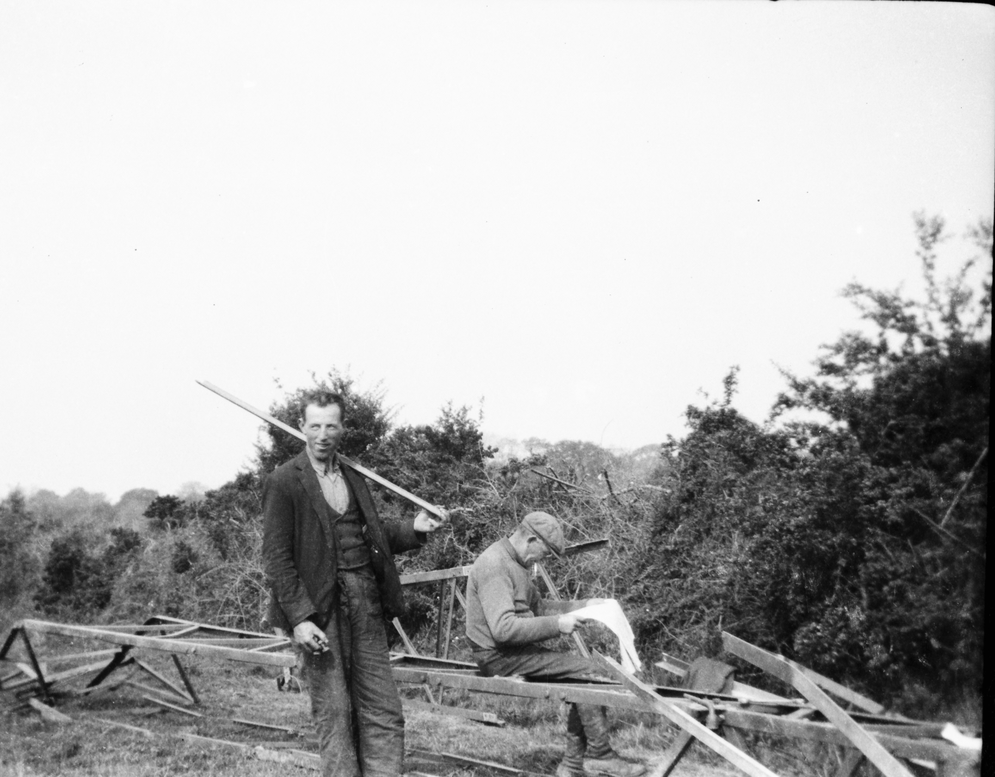Two workers among framework sections laid out on the ground, with one holding a long timber pole and the other reviewing a document beside hedgerows in the countryside.