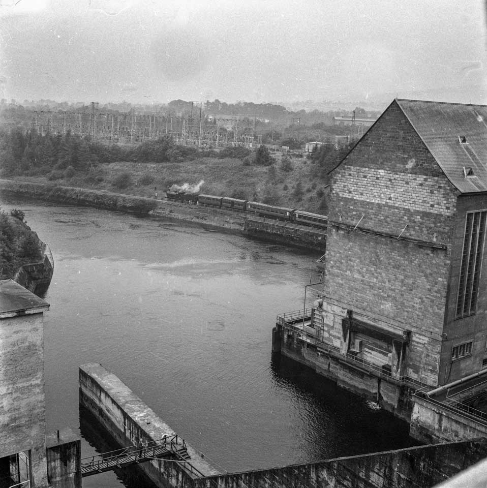Black-and-white photograph showing a steam excursion train travelling along a railway line beside the river, with hydroelectric station buildings and electrical infrastructure visible in the foreground and background.