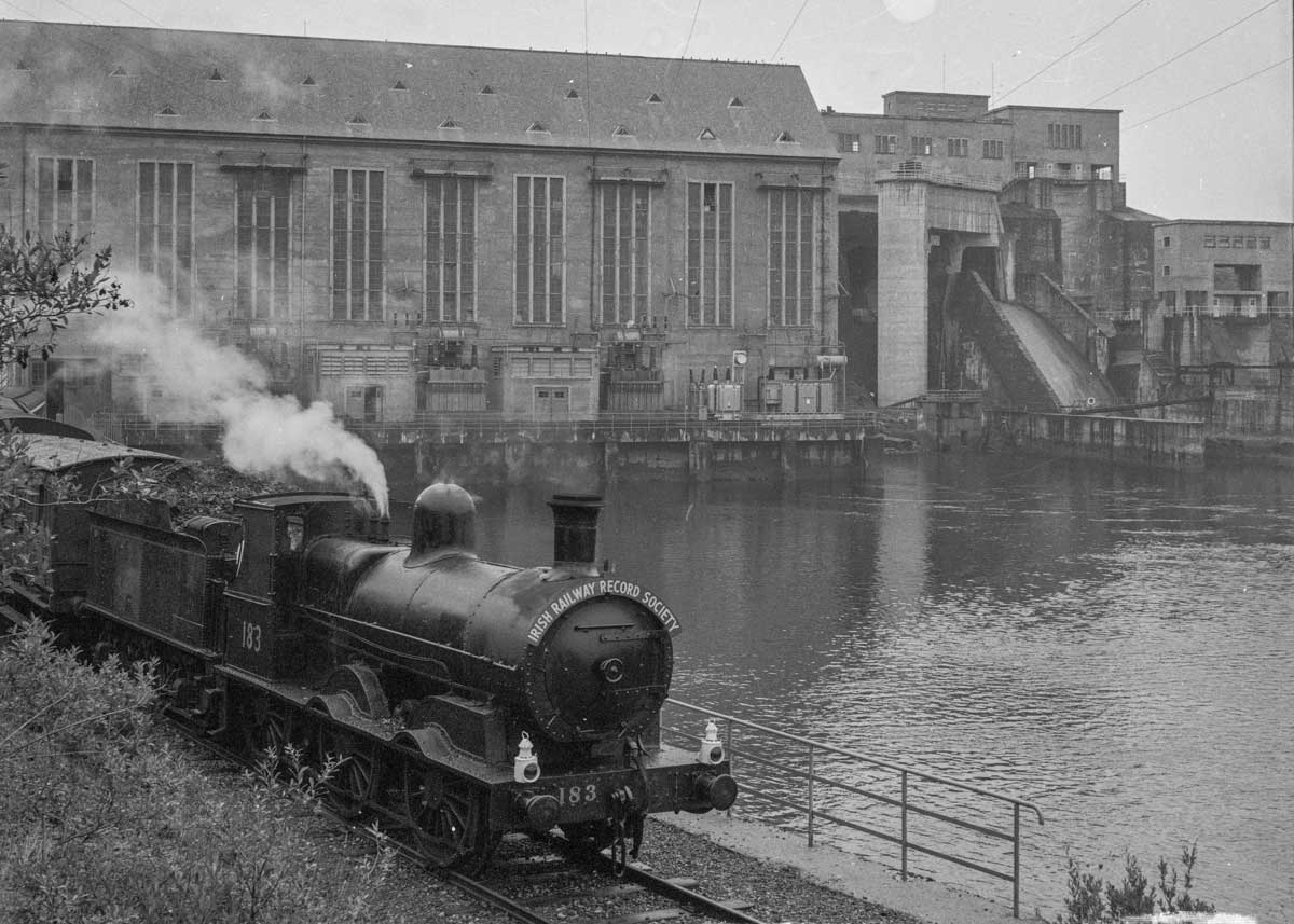 black-and-white photograph showing a steam excursion train travelling beside the hydroelectric power station, with steam rising from the locomotive as it passes along the waterside track.