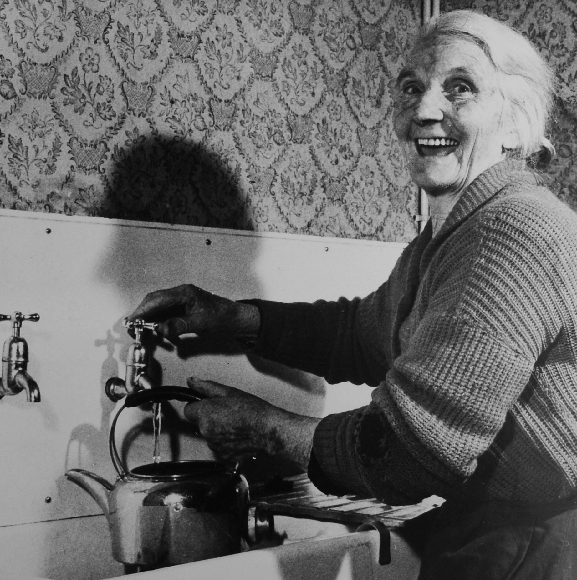 Elderly woman beaming as she turns on a tap to fill a kettle, 1940s.