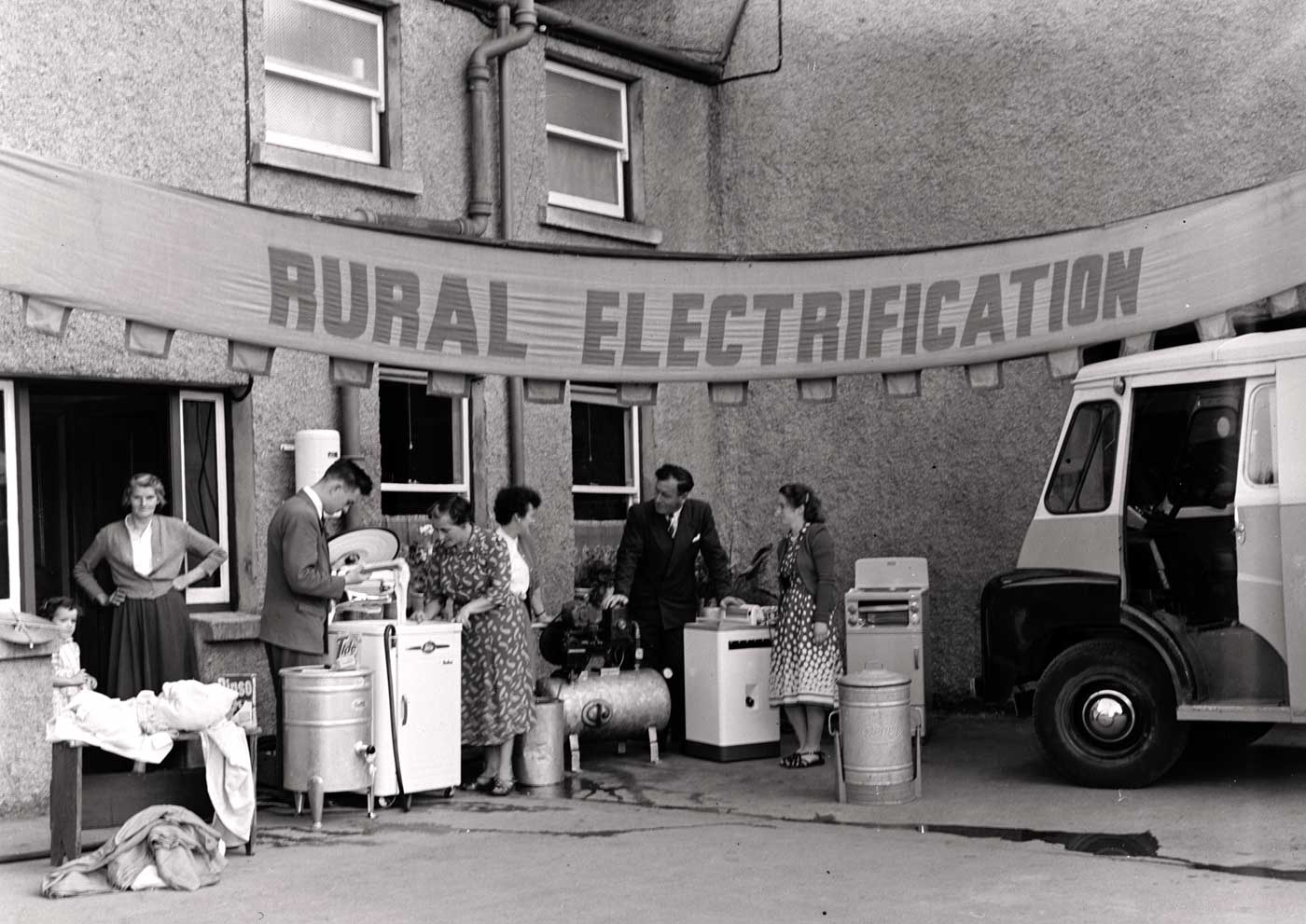 Black-and-white photograph showing a rural electrification demonstration outside a building. A banner reading “RURAL ELECTRIFICATION” hangs overhead while several people stand among household electrical appliances and equipment beside a utility vehicle.
