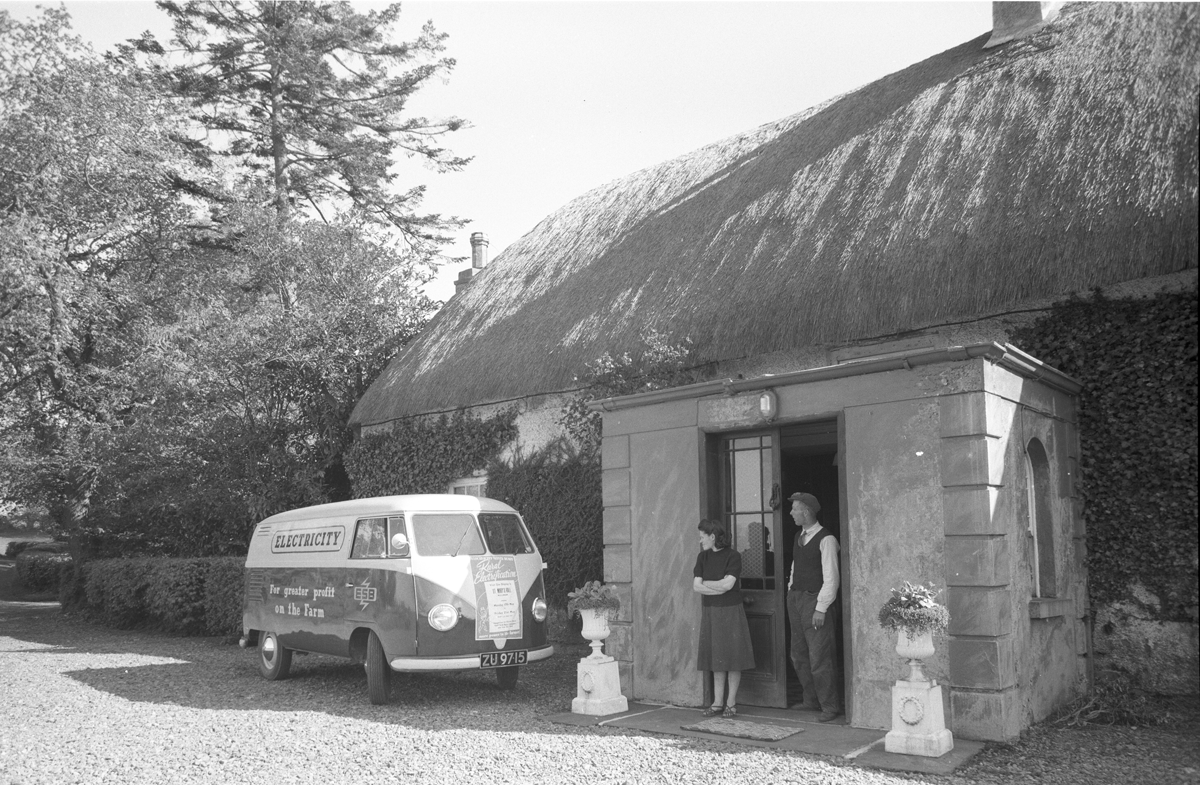 Black-and-white photograph showing an ESB promotional van parked outside a thatched farmhouse, with two people standing in the doorway.