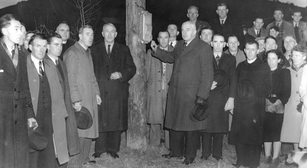 Black-and-white photograph showing a crowd gathered around a wooden utility pole during a rural electrification switching-on ceremony, with a person operating an electrical switch mounted on the pole.