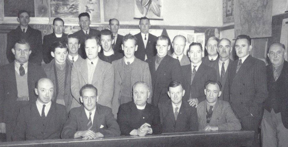 Black-and-white group photograph showing a committee meeting, with several people seated at a table in the front row and others standing behind them in a meeting room.