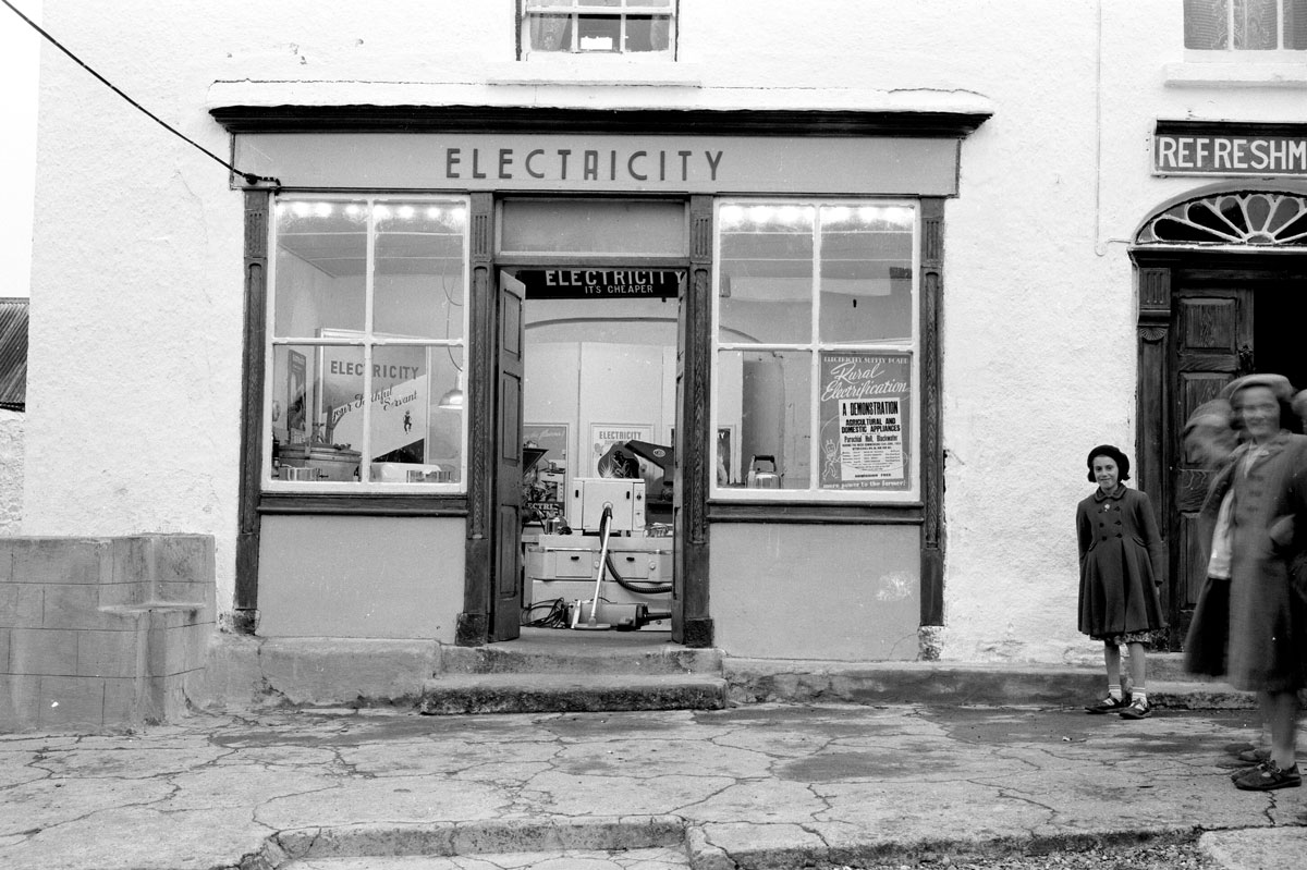 Black-and-white photograph showing a shopfront used for a pop up electrical appliance showroom, with electrical appliances displayed inside and promotional signs visible in the windows.