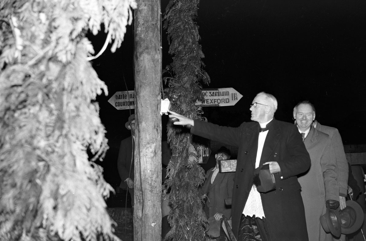 Black-and-white photograph showing a ceremonial switching-on event, with a person activating an electrical switch mounted on a decorated wooden pole while onlookers stand nearby.