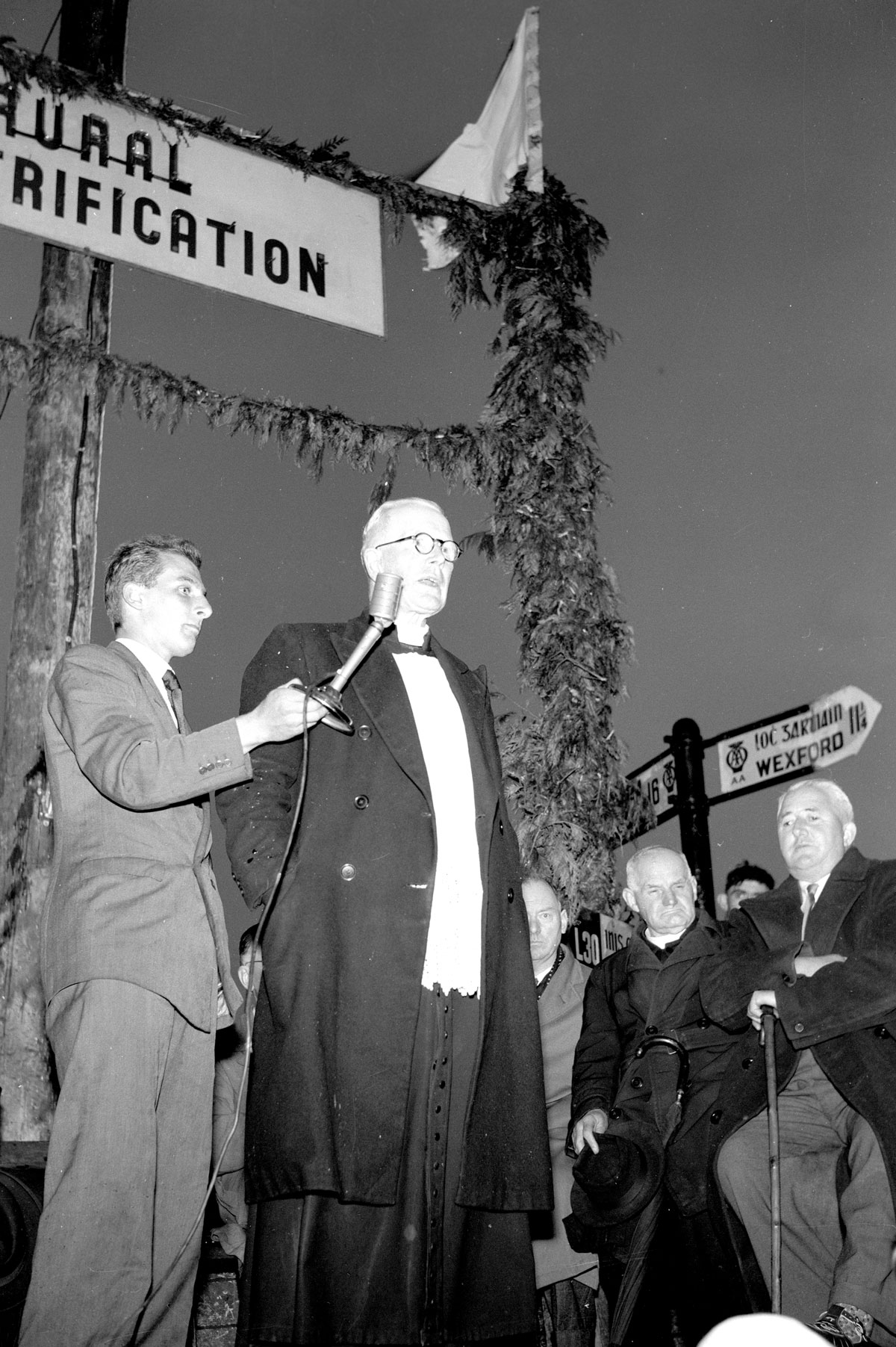 Black-and-white photograph showing a public ceremony beneath a rural electrification banner, with a speaker addressing a crowd using a microphone.