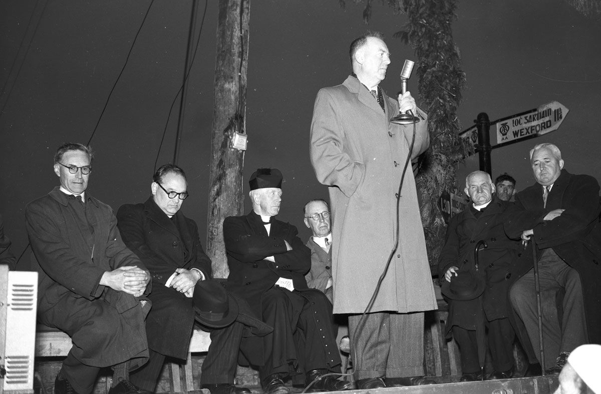 Black-and-white photograph showing a speaker addressing an outdoor gathering from a raised platform, using a microphone while several attendees are seated nearby.
