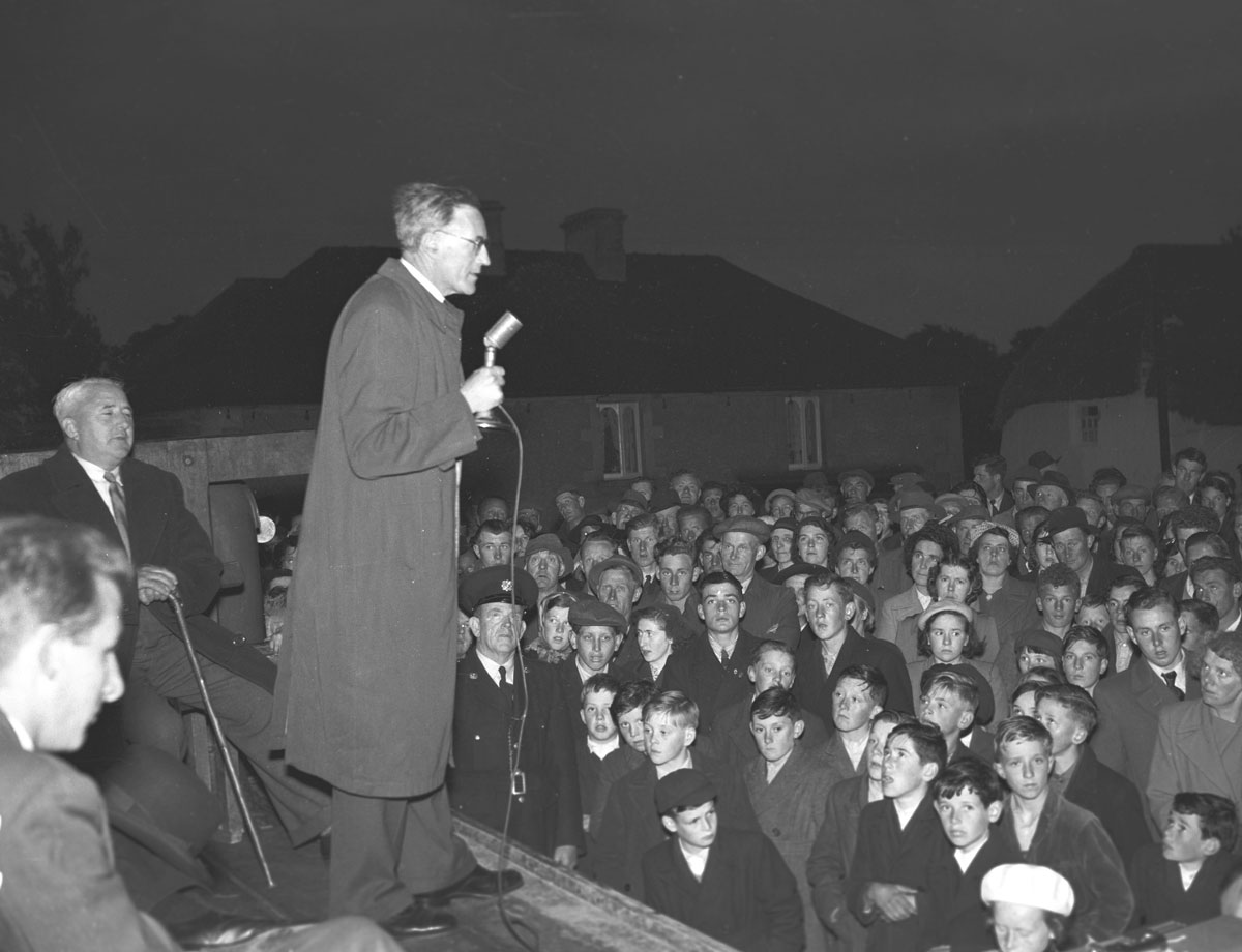 Black-and-white photograph showing a speaker addressing a large crowd at an outdoor evening gathering, using a microphone from a raised platform.