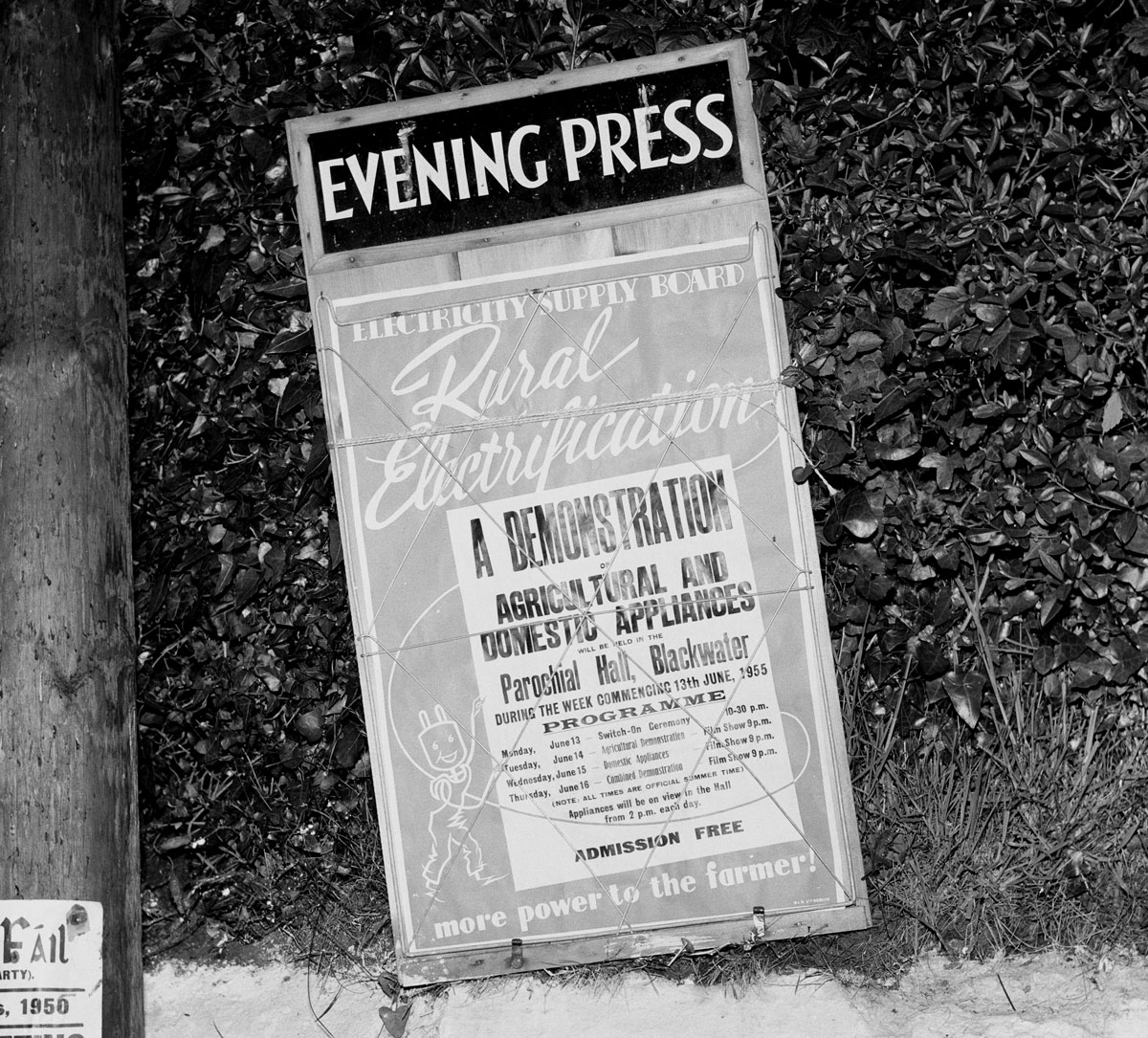 Black-and-white photograph of a roadside advertising sign promoting a rural electrification demonstration of agricultural and domestic electrical appliances.