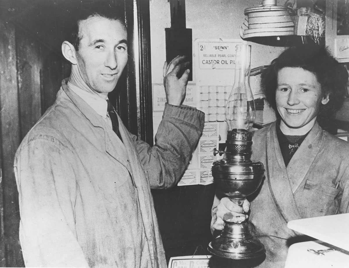 Black-and-white photograph showing two people indoors beside a paraffin lamp, with one person operating an electrical switch mounted on a wall.