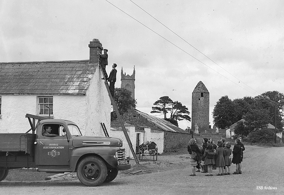 Black-and-white photograph showing rural electrification work at a house, with two workers on ladders installing electrical equipment while local children watch. An ESB lorry is parked beside the building.