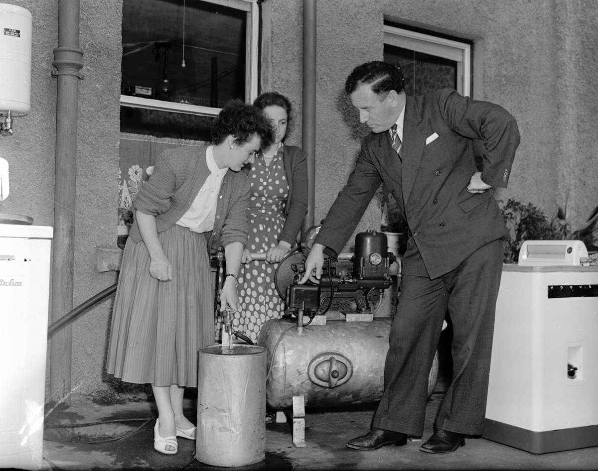 Black-and-white photograph showing a demonstration of an electrically powered water pump system, with three people examining the pump and water flowing into a metal container.