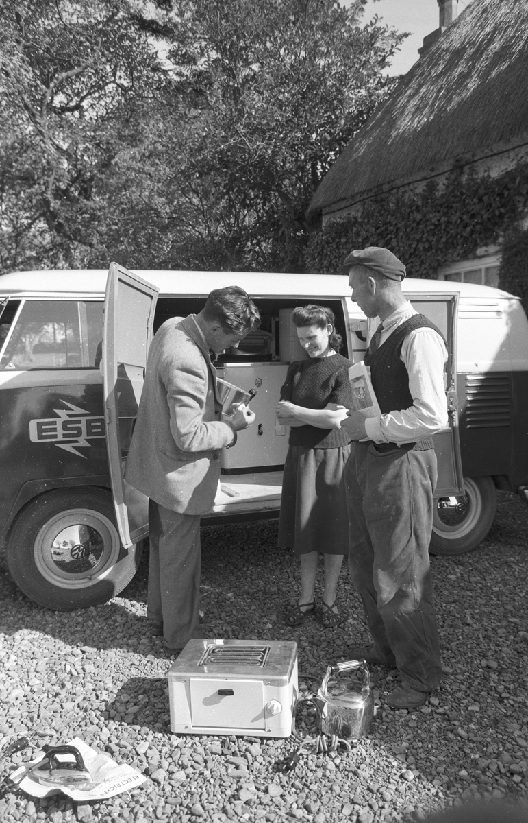 Black-and-white photograph showing an ESB representative demonstrating electrical appliances from a promotional van to two people outside a thatched farmhouse.