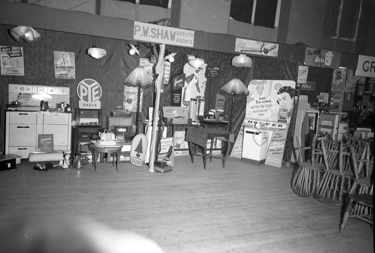 Black-and-white photograph showing a display of domestic electrical appliances, radios and household equipment at an indoor exhibition.