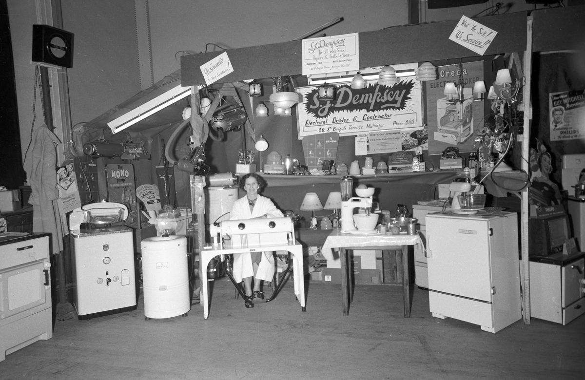 Black-and-white photograph showing a domestic appliance exhibition stand, with a demonstrator seated among electrical appliances, lighting fixtures and household equipment.