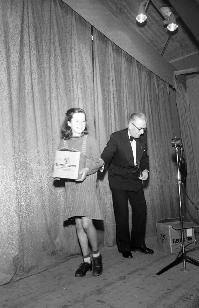 Black-and-white photograph showing a prize presentation on a stage, with a child receiving an electric toaster from a presenter.