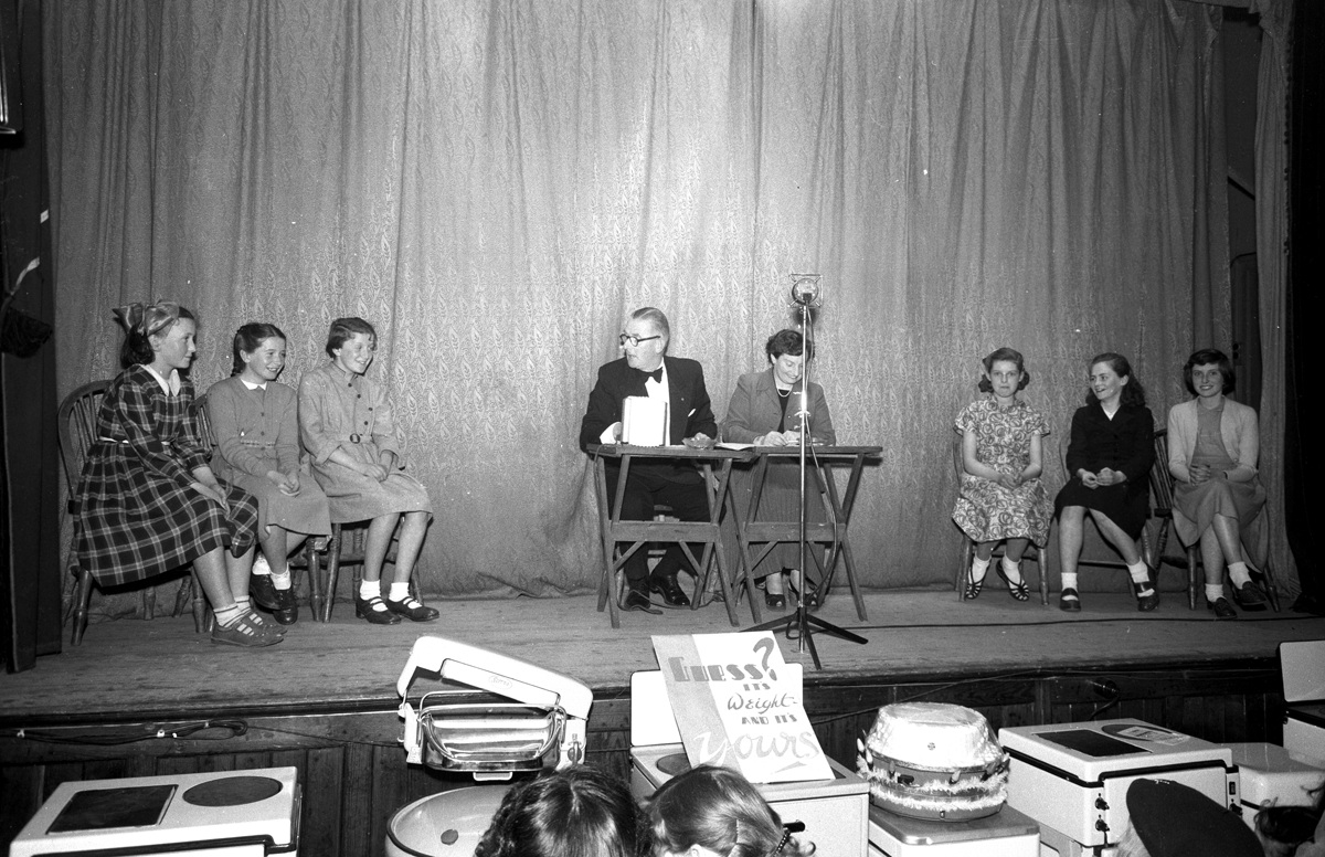 Black-and-white photograph showing a panel discussion or quiz on a stage, with participants seated on either side of a table and electrical appliances displayed in the foreground.