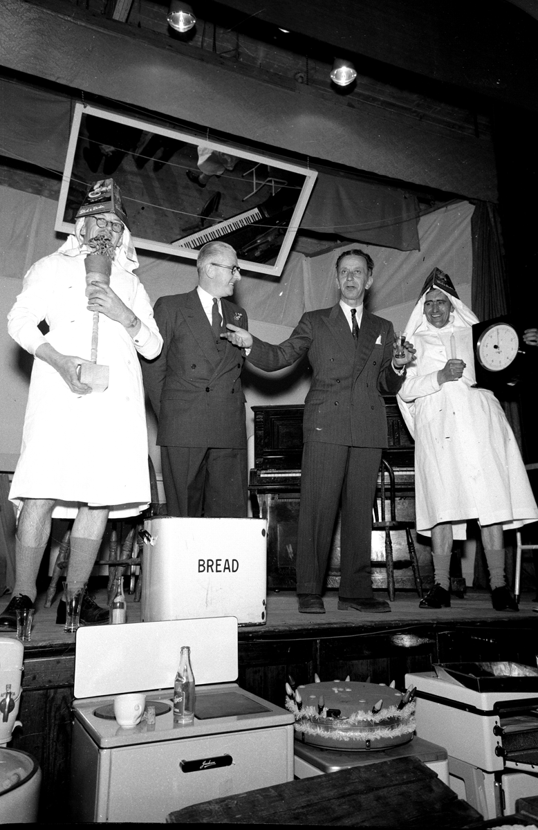 Black-and-white photograph showing a stage performance or community entertainment event, with costumed participants standing among household electrical appliances displayed as prizes.