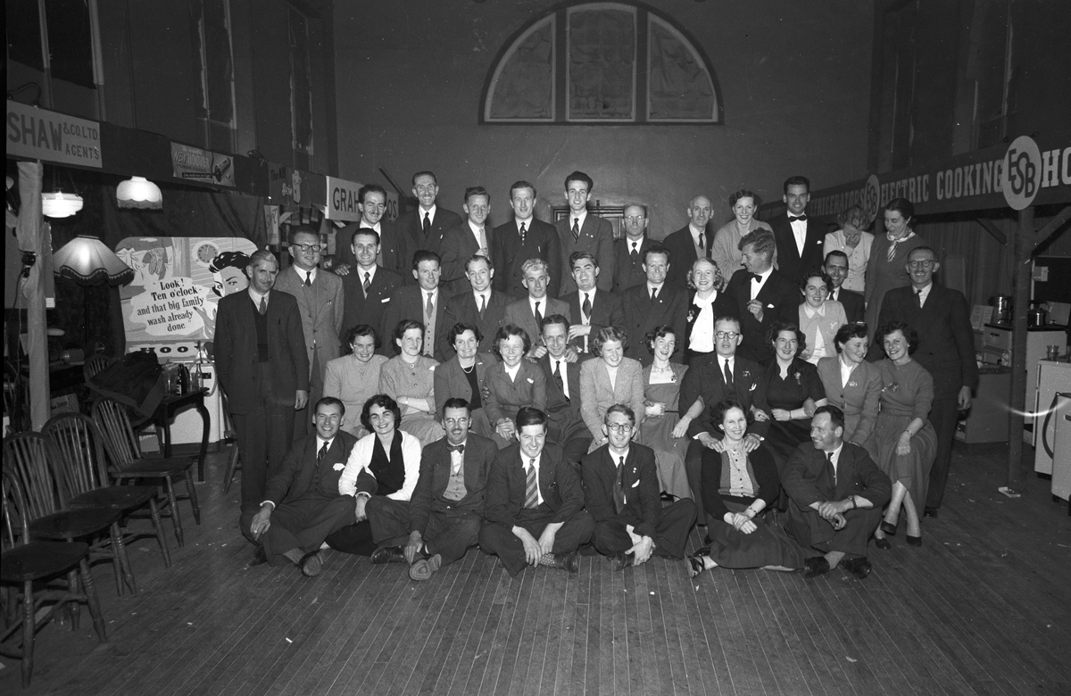 Black-and-white group photograph showing attendees gathered inside an exhibition hall, with displays of household electrical appliances visible around them.