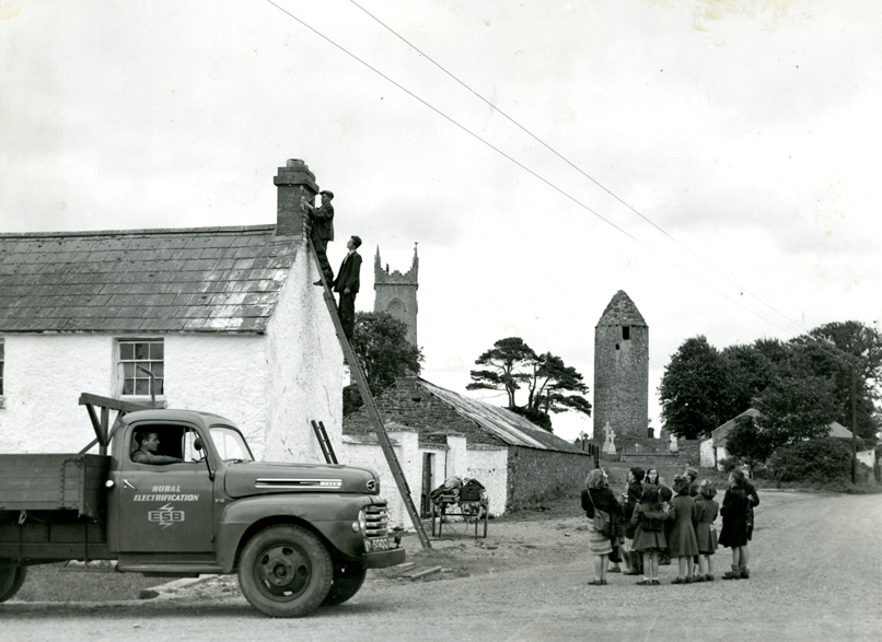 Two men stand atop a ladder leaning against a cottage while children look on